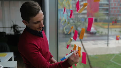 Smiling-handsome-young-man-writing-on-sticky-notes-on-window.